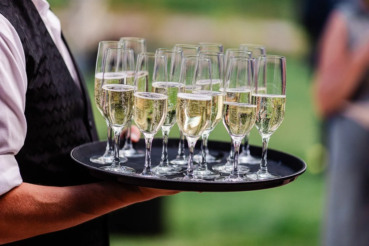 A waiter in a black vest carries a tray of filled champagne flutes. The setting is outdoors, with a blurred green background, suggesting a celebratory event.