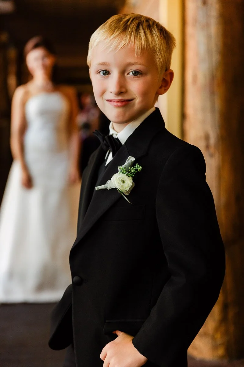 A young boy in a black tuxedo with a white boutonniere smiles confidently. Behind him, a woman in a white dress stands blurred in the background.