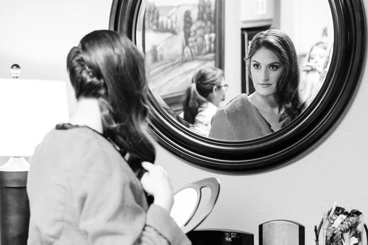 A woman gazes into a round mirror, adjusting her hair with focused expression. Soft lighting and a serene room create a contemplative mood.