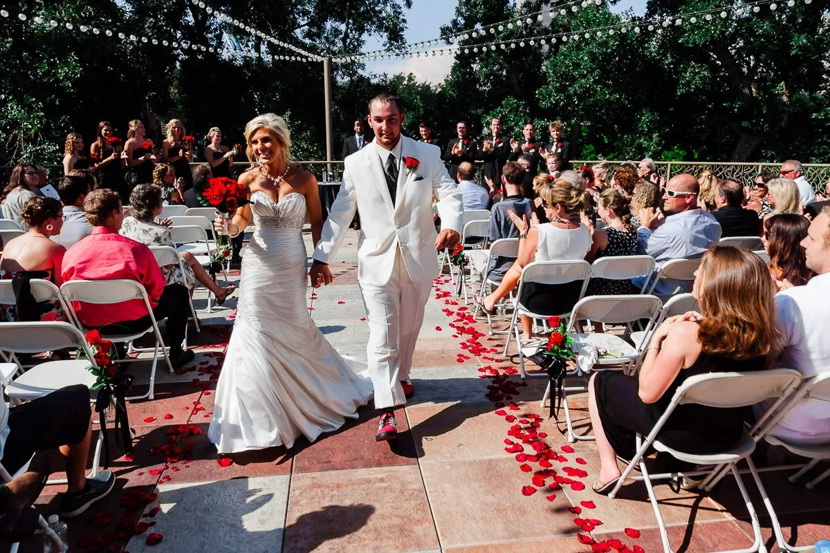 Bride in a white gown and groom in a white suit walk down an aisle strewn with red rose petals at a Lincoln Center wedding in Fort Collins. Guests seated on both sides clap under string lights.