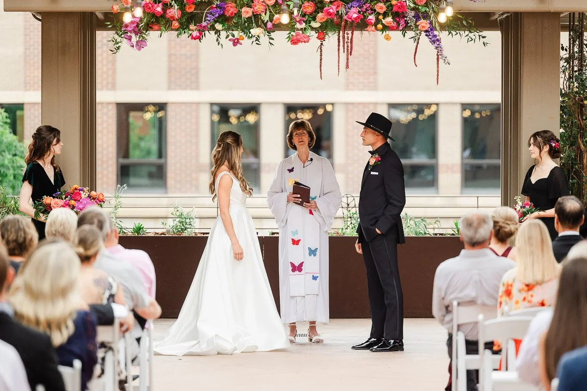 A bride and groom stand under a vibrant floral arch during a wedding ceremony. The officiant, in white with butterfly patterns, is speaking. Guests are seated, watching attentively.