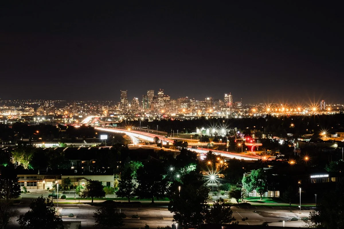 A vibrant cityscape at night with bright lights illuminating the skyline. A busy highway with blurred car lights leads into the city, evoking a lively, bustling atmosphere.