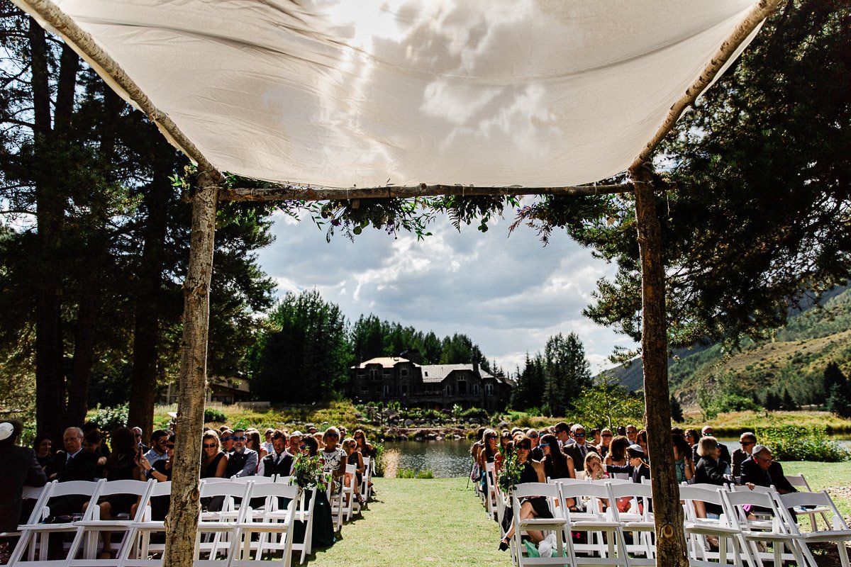 Outdoor wedding with guests seated on white chairs under a canopy. The backdrop features a scenic lake, lush trees, and a large house, creating a serene atmosphere.