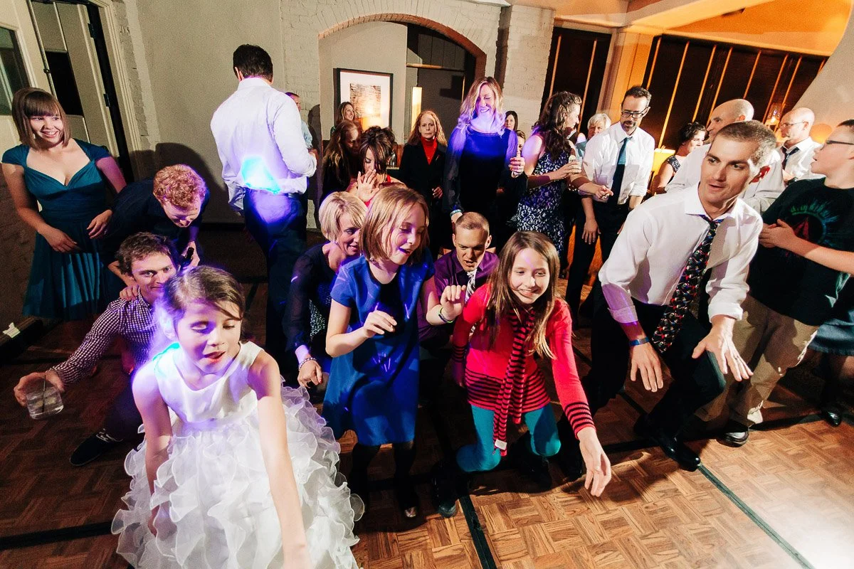 A lively dance floor scene with adults and children wearing formal attire, joyfully dancing. The mood is festive and energetic, suggesting a celebration.