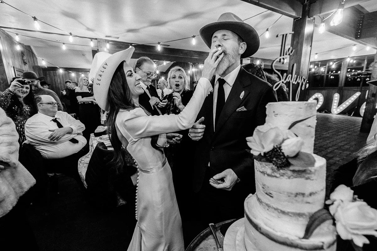 Bride in a white hat playfully feeds cake to a laughing groom in a suit and hat at a lively, warmly-lit celebration, with guests smiling and cheering.