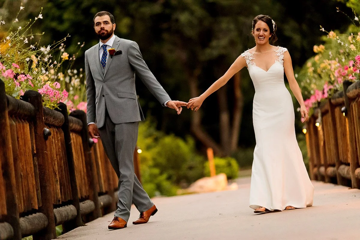 A bride in a white gown and a groom in a gray suit hold hands while walking on a flower-lined wooden bridge. They appear happy and relaxed during their Chatfield Farms wedding in Littleton, Colorado