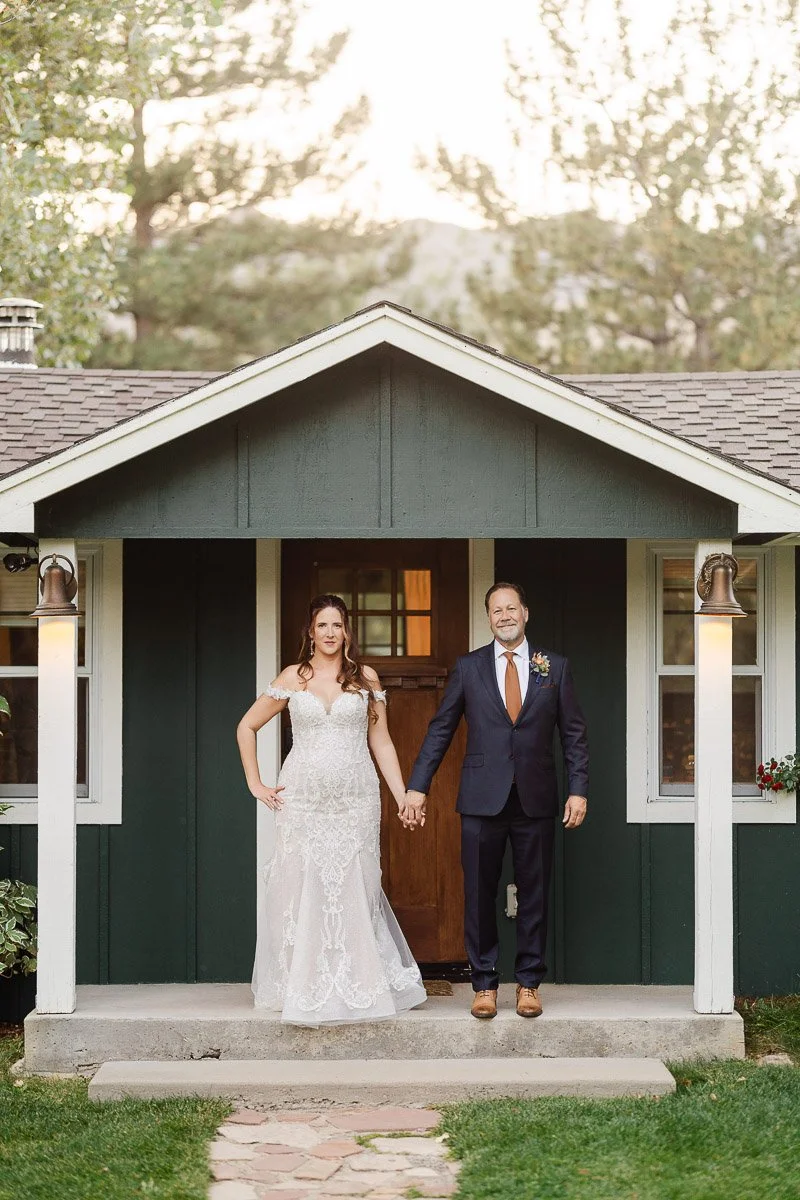 A bride in a lace gown and a groom in a navy suit stand holding hands, smiling in front of a small green cottage with white trim, set in a forested area.