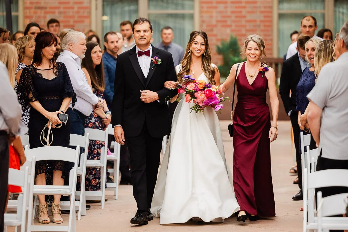 Bride in white gown with vibrant bouquet walks down aisle with parents. They smile warmly at rows of seated guests in an outdoor ceremony.
