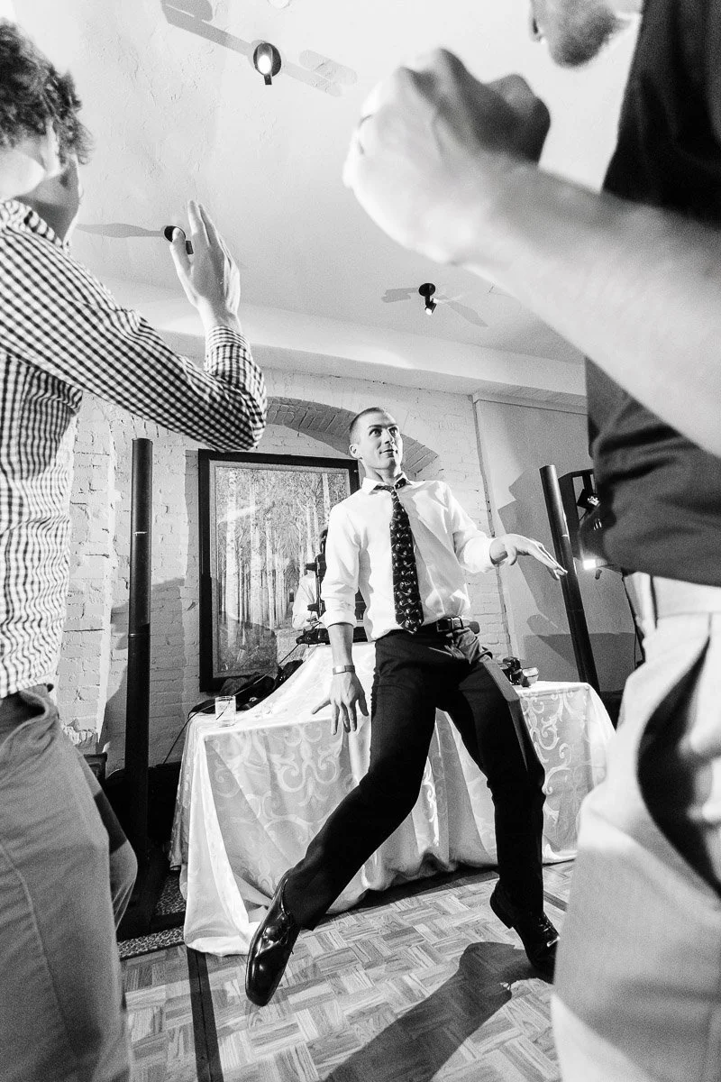 A man energetically dances in a room, wearing a shirt and tie, surrounded by people. The scene is lively and joyful, set in a decorated venue.