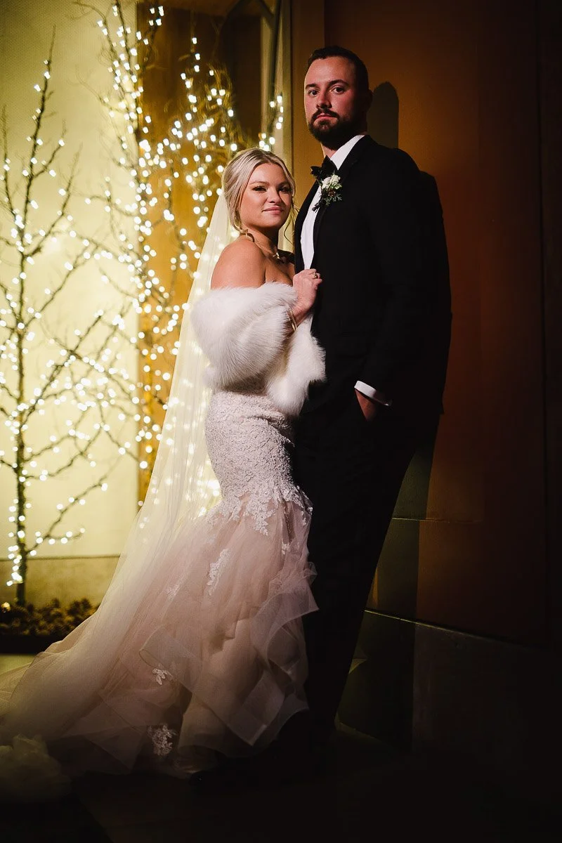 A couple in wedding attire poses romantically indoors. The bride wears a lace gown and white wrap, while the groom is in a black suit. Soft lights twinkle on a bare tree in the background, creating an intimate, warm ambiance.