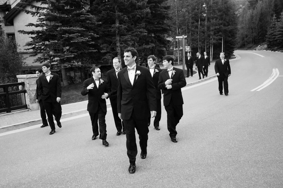 A group of groomsmen in tuxedos walk confidently down a curved road lined with trees, portraying a sense of camaraderie and anticipation.
