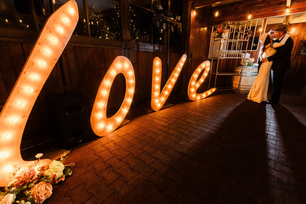 A couple shares a romantic embrace in a warmly lit room. Illuminated "Love" letters and a bouquet enhance the cozy, festive atmosphere captured by La Joya Dulce wedding photographer tomKphoto