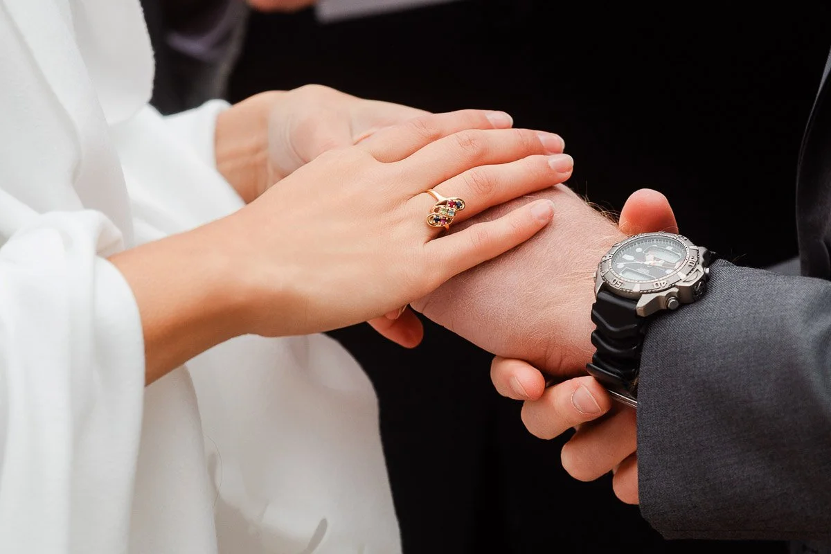 Close-up of a couple holding hands; a woman wears a ring, a man a watch. The woman's hand gently rests atop the man's, evoking a feeling of tenderness.