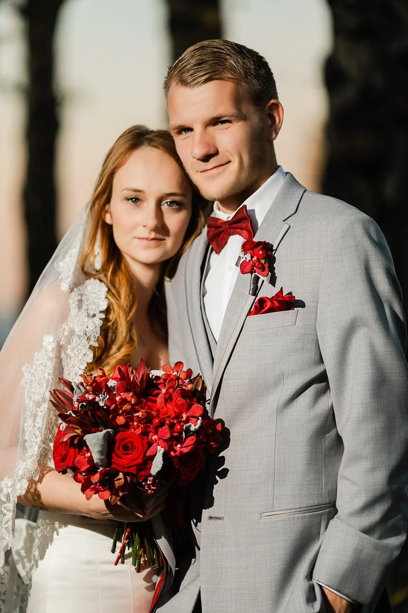 Bride in a lace veil holds a vibrant red bouquet, standing closely beside the groom in a gray suit with matching red accents, under a warm light.