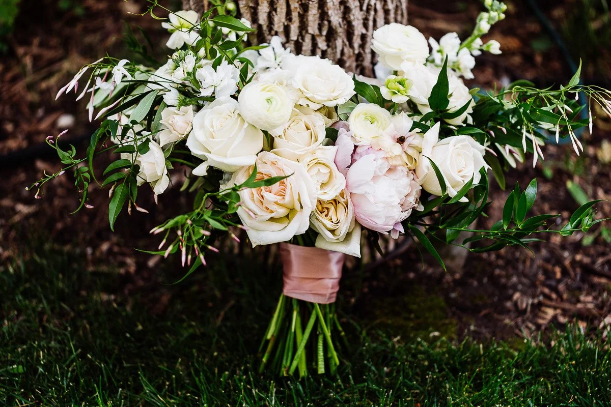 A bridal bouquet featuring white and blush roses with lush greenery, wrapped in pink ribbon, rests against a tree trunk on a grassy lawn. Elegantly romantic.