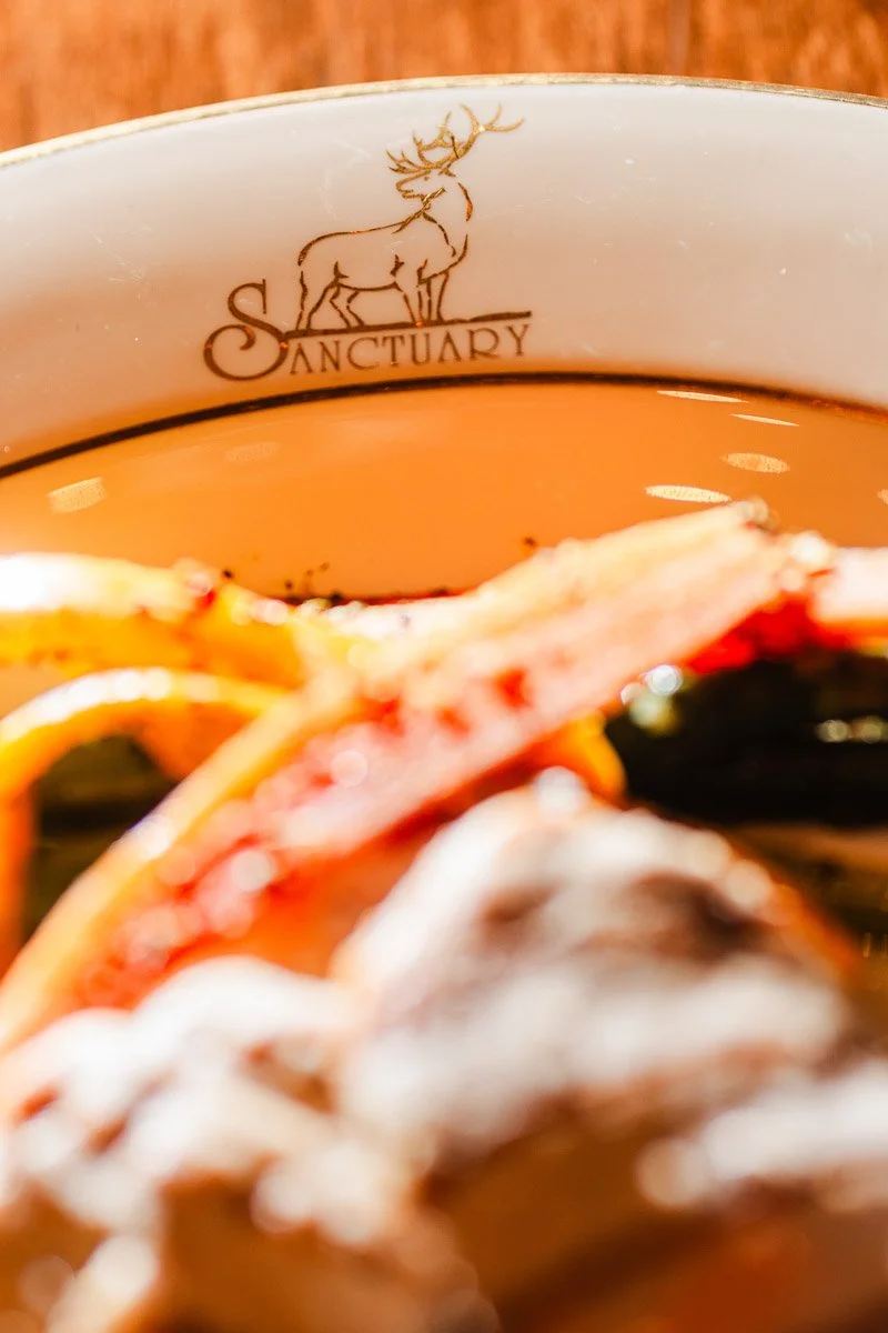 Close-up shot of a decorative plate with "Sanctuary" and a deer emblem. Blurred colorful food items in the foreground create a warm, inviting atmosphere.