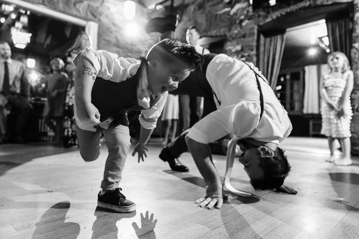 Black and white photo of a young boy and a man dancing energetically on a wooden floor, surrounded by a cheerful crowd in a warmly lit room.