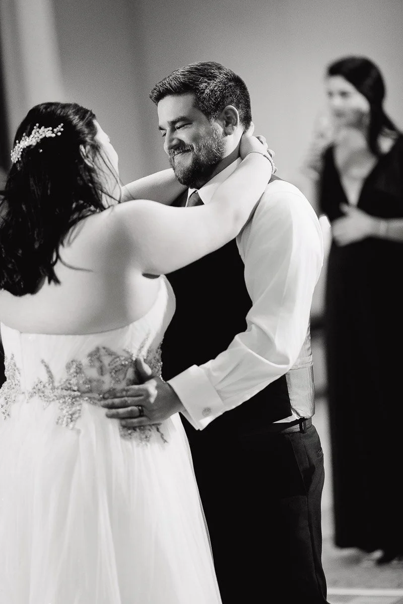 A couple dances closely, smiling at each other in a warmly lit room. The bride wears a beaded dress, and the groom wears a vest. A woman watches warmly in the background.