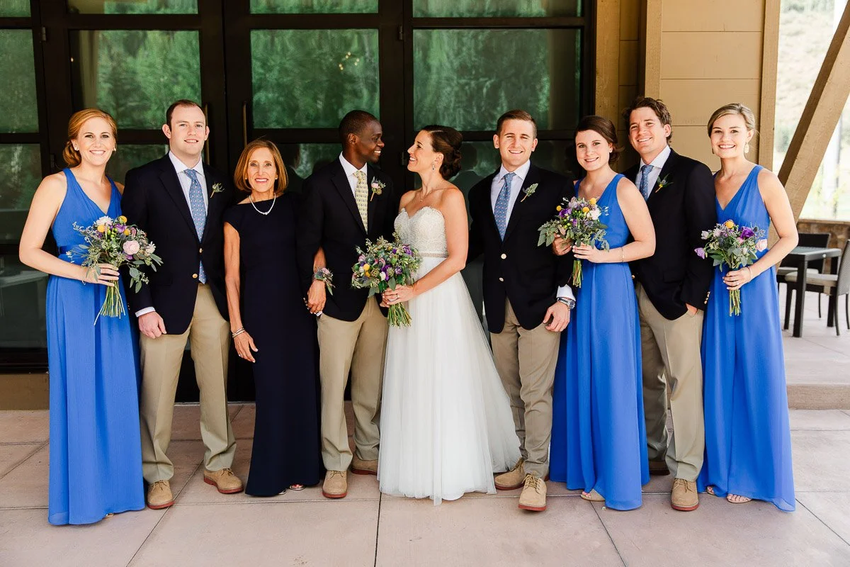 A joyful wedding party poses. The bride, in a white gown, is surrounded by bridesmaids in blue dresses and groomsmen in suits. All smile warmly.