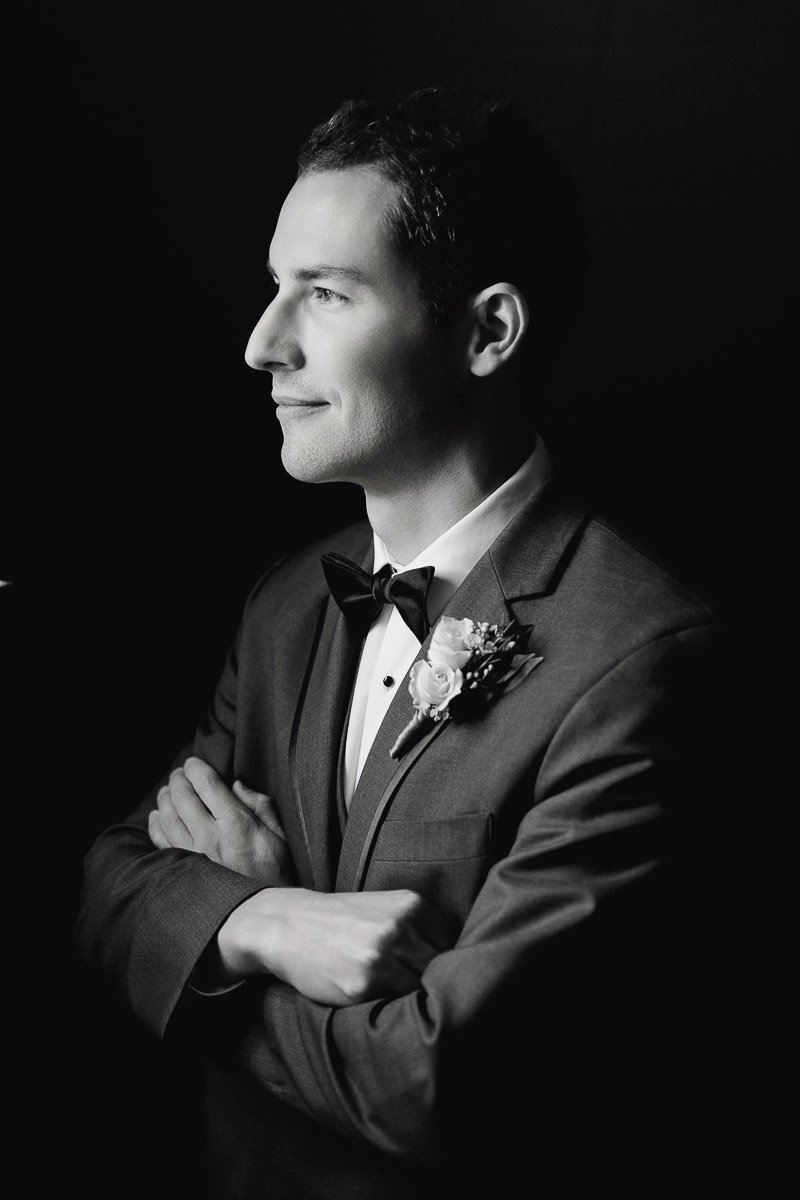 Black and white portrait of a man in a suit with a bowtie and floral boutonniere, arms crossed, looking confident and thoughtful to the left.