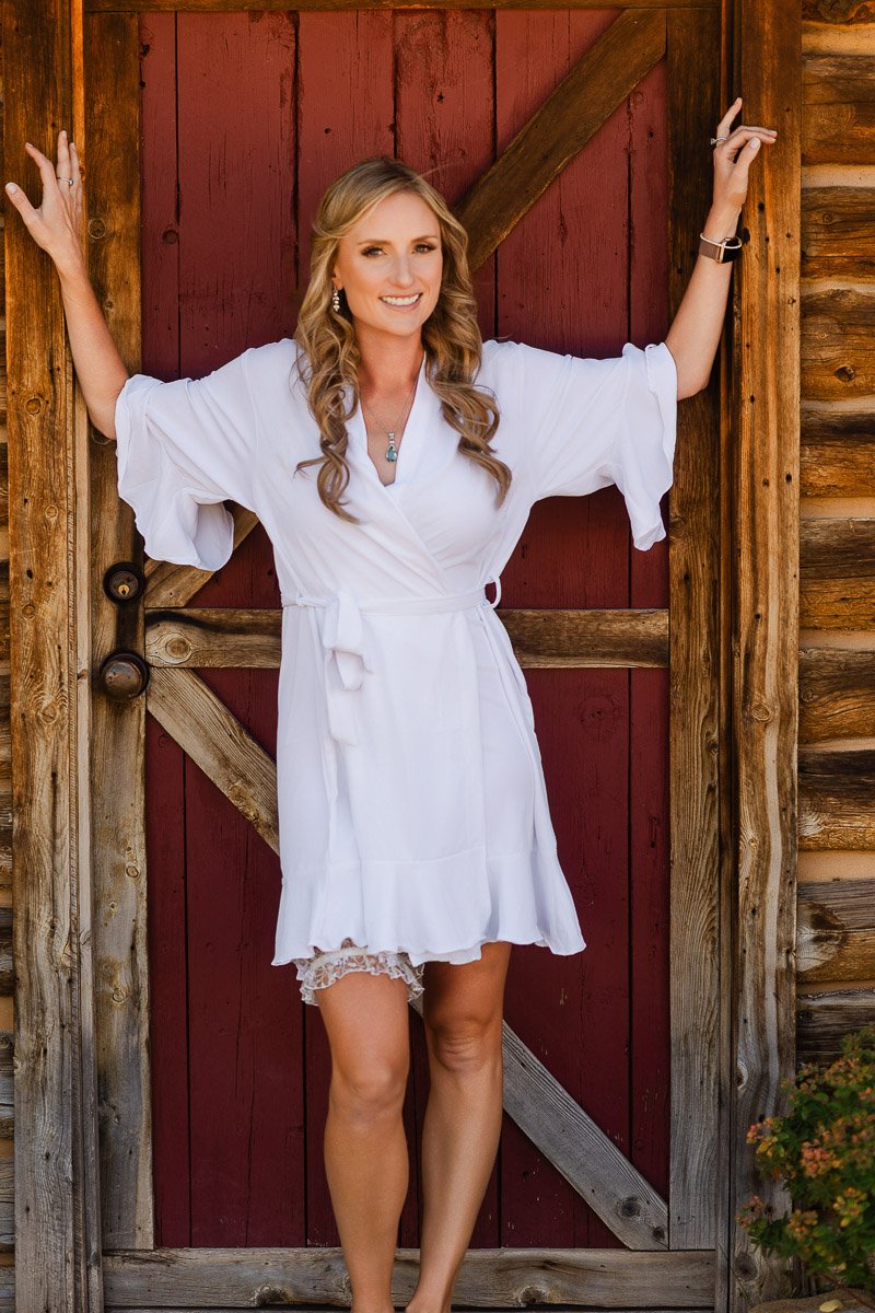 Smiling woman in a white dress stands in front of a rustic wooden door with burgundy panels, exuding a relaxed and cheerful vibe.
