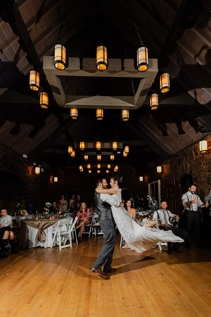 A couple is dancing joyfully in a warmly lit hall with high wooden ceilings and hanging lanterns. Guests sit and watch, smiling, creating a festive ambiance.