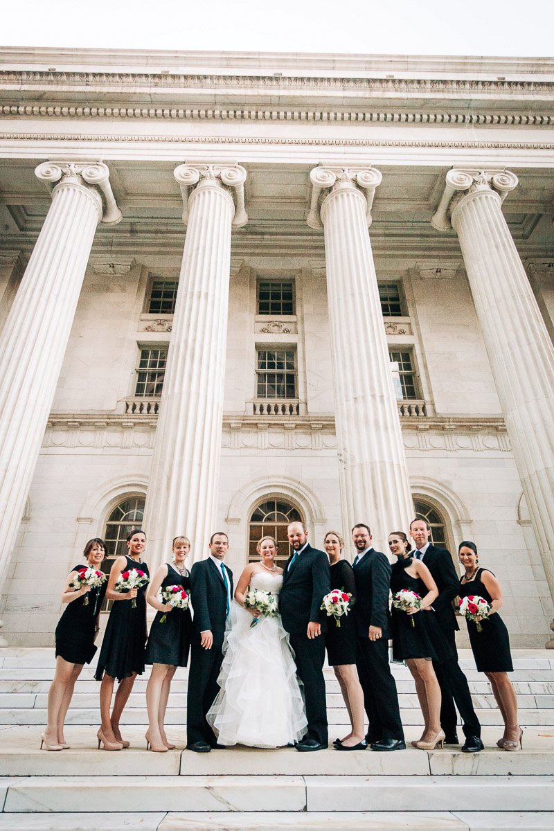A wedding party poses on steps in front of a grand building with tall columns. The bride and groom are centered, surrounded by bridesmaids in black dresses holding bouquets. The tone is elegant and celebratory.