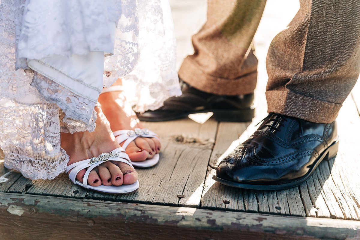 Close-up of a bride's lace dress and sandals next to a groom's brown suit pants and polished black dress shoes on a sunlit wooden floor captured by YMCA of the rockies wedding photographer tomKphoto