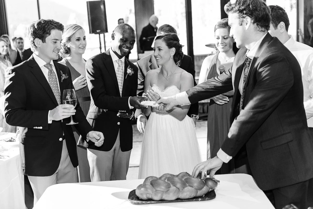 A joyful group, including a bride in a strapless gown and a groom in a suit, gather around a loaf of challah bread, engaging in a wedding tradition.