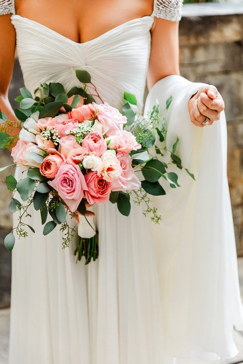 A bride in an elegant white dress holds a bouquet of pink and peach roses with greenery, exuding romance and joy against a stone wall backdrop.