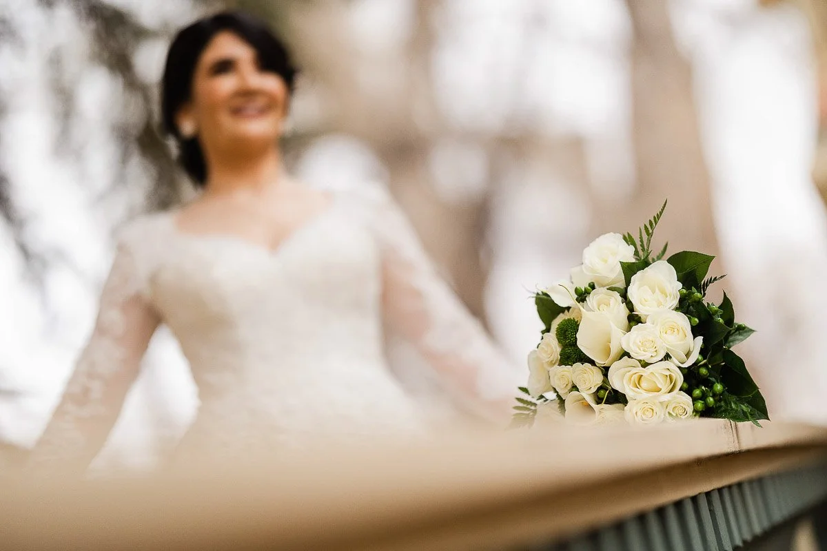 A bride in a lace wedding dress stands in the background, smiling. In the foreground, a bouquet of white roses and greenery rests on a wooden railing.