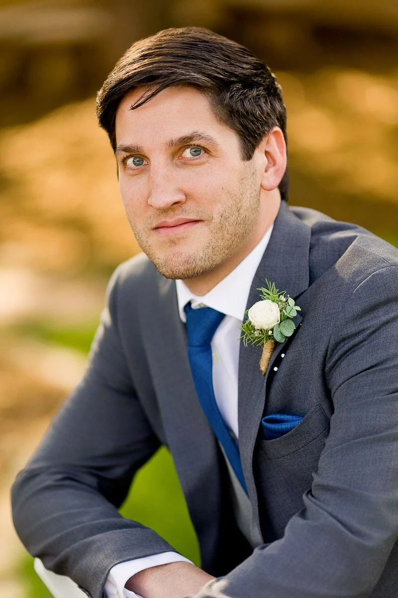 Groom in a gray suit with blue tie, adorned with a white boutonniere, gazes thoughtfully. Background is softly blurred, suggesting a serene setting.