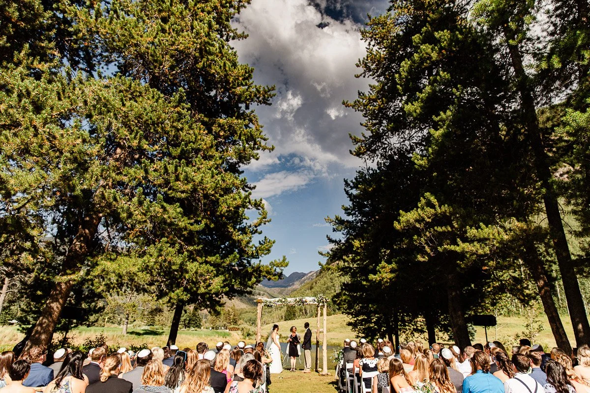 Outdoor wedding ceremony under a floral arch amidst tall trees, with guests seated on either side, set against a backdrop of mountains and a blue sky during a Vail Golf Club wedding in Vail, Colorado