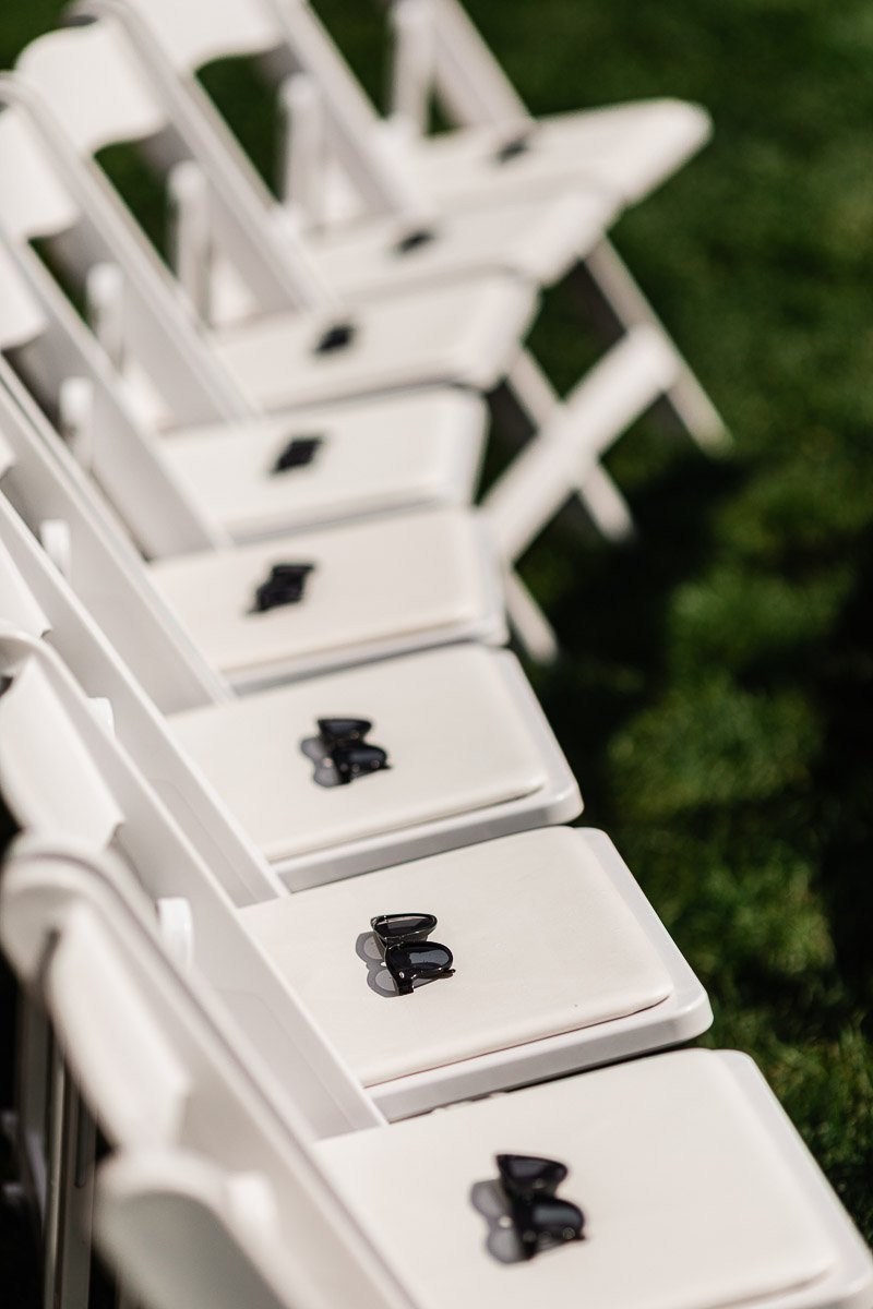 Rows of white folding chairs on grass, each with a pair of black sunglasses placed on the seat, suggesting an outdoor event or ceremony setup.