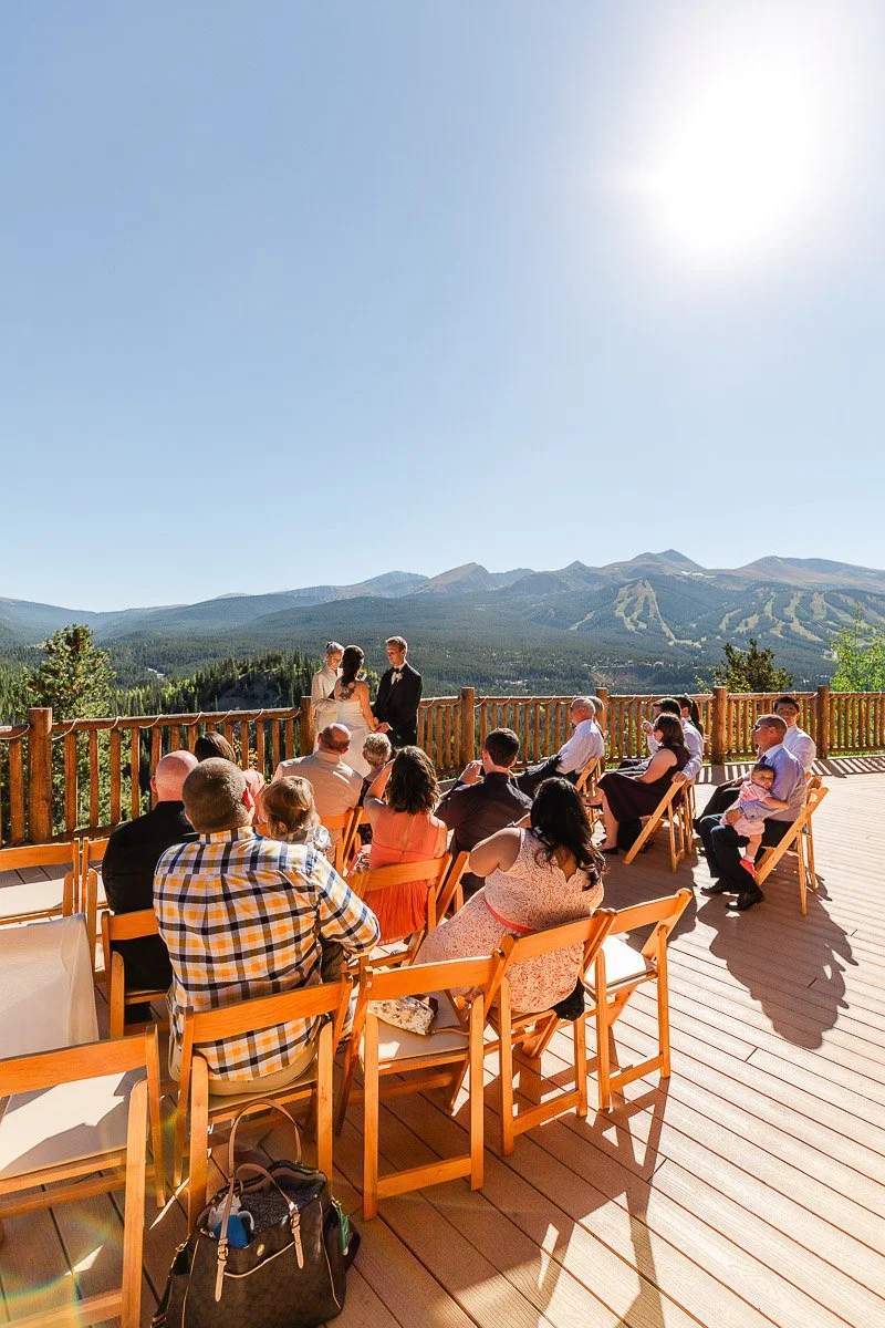 Outdoor wedding ceremony on a sunny deck with mountain views. Guests sit in wooden chairs, facing the couple and officiant, conveying a joyful, warm atmosphere.