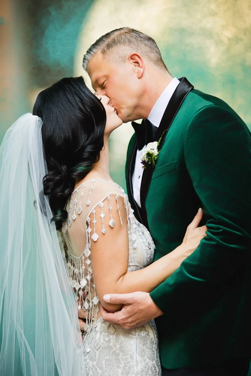 A bride and groom share a kiss, the bride in a lace dress with a veil, and the groom in a green velvet jacket. The scene is romantic and intimate.
