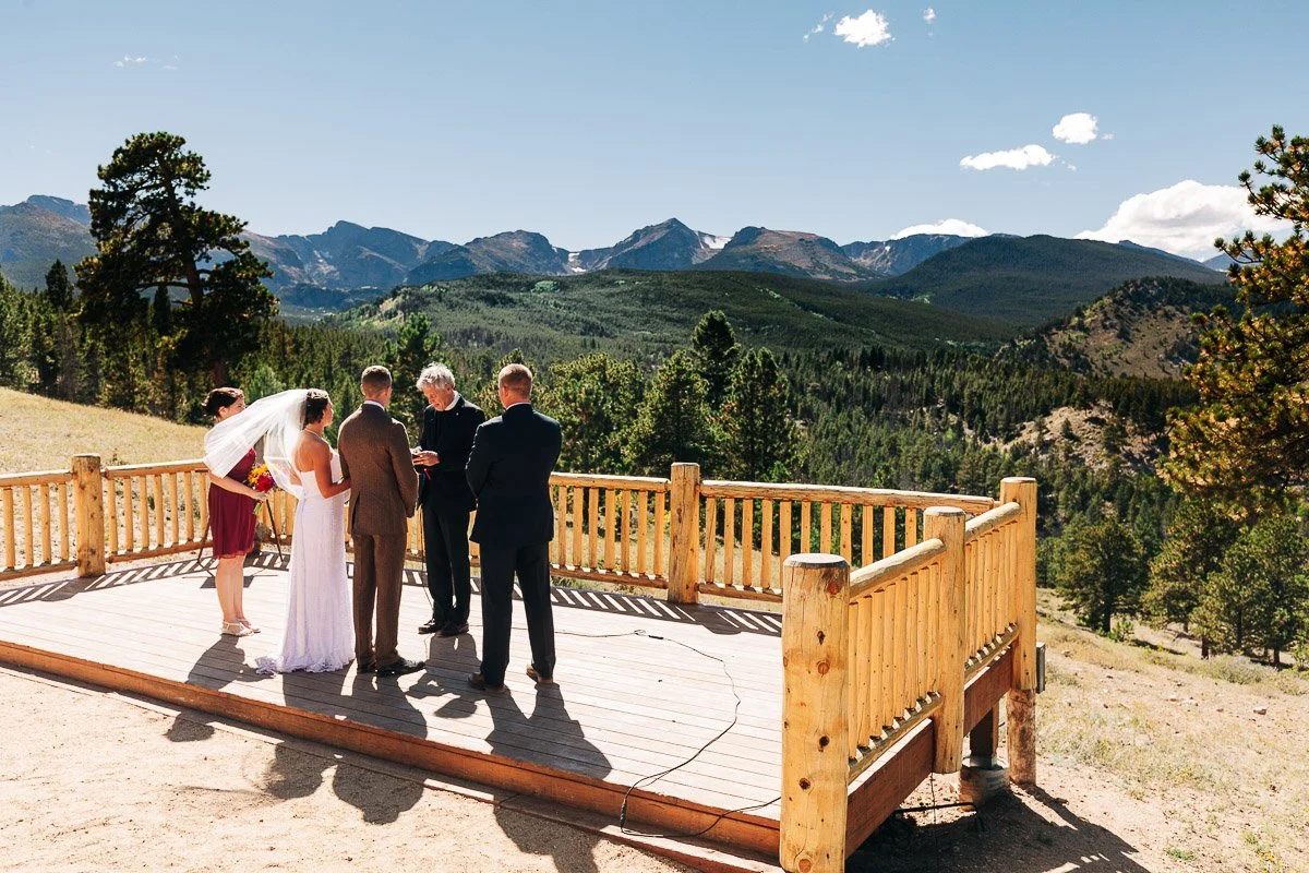 A couple stands facing each other during their outdoor wedding on a wooden deck, surrounded by mountains and trees under a clear blue sky.