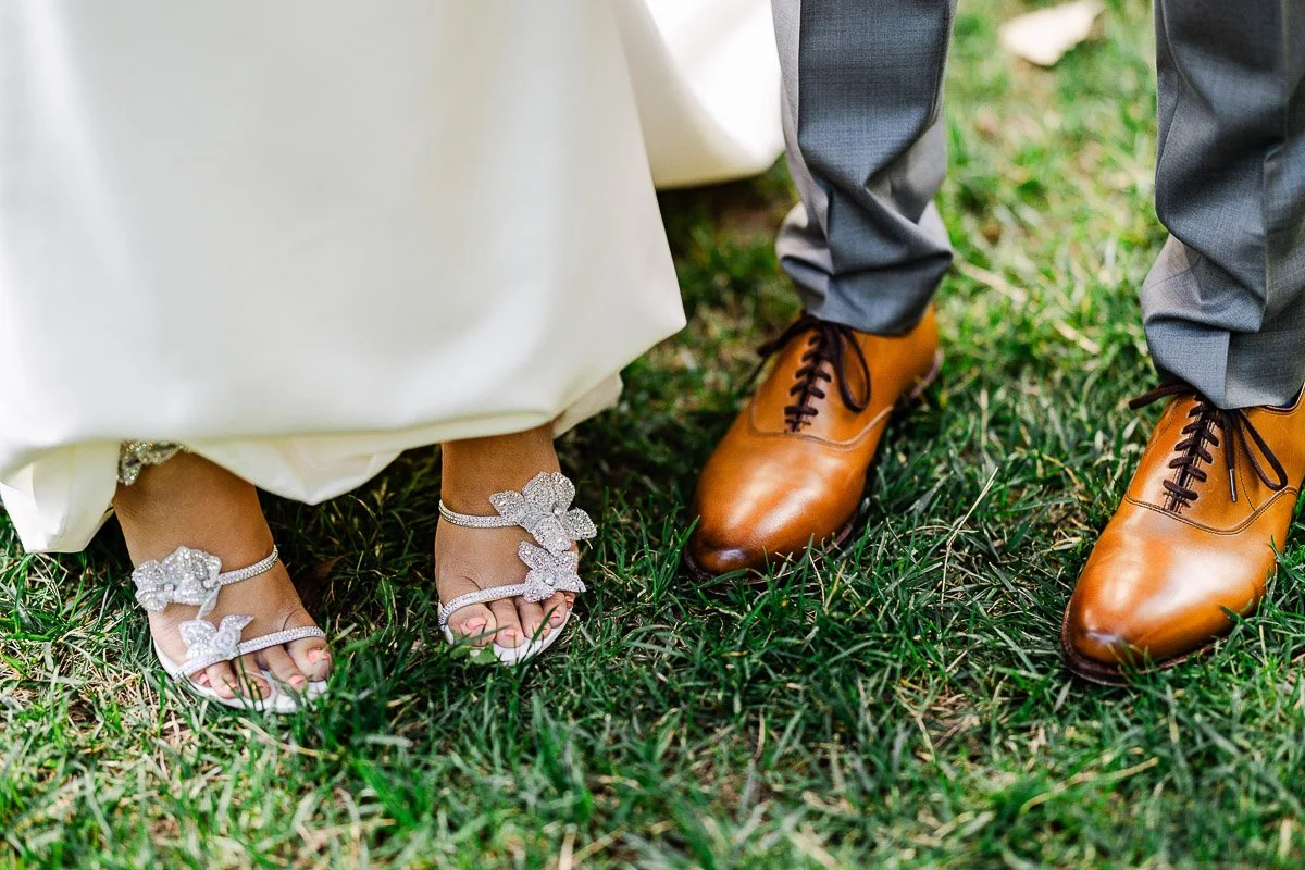 Close-up of a bride and groom's footwear on grass. The bride wears sparkly sandals; the groom wears tan leather shoes. Elegant, celebratory mood.