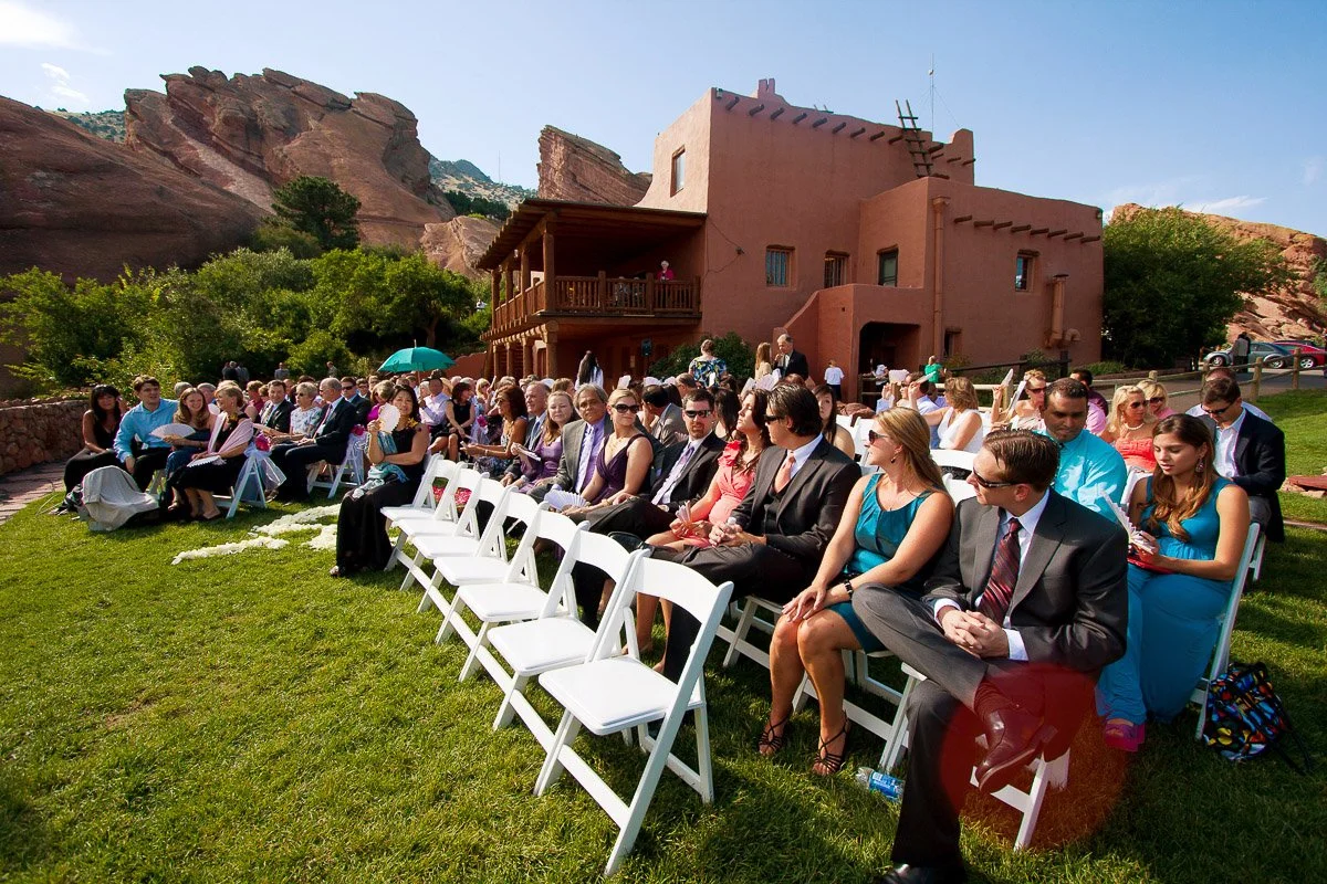 Outdoor wedding ceremony with guests seated on white chairs in bright sunlight. Rustic adobe building and red rock formations in the background.