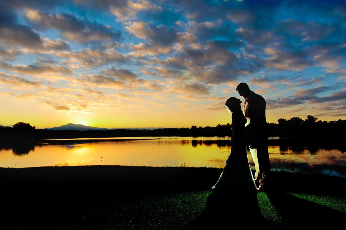 A couple stands silhouetted against a vibrant sunset by a tranquil lake, with colorful clouds reflecting on the water. The scene feels romantic and serene during a Barn at Raccoon Creek wedding in Littleton, Colorado.