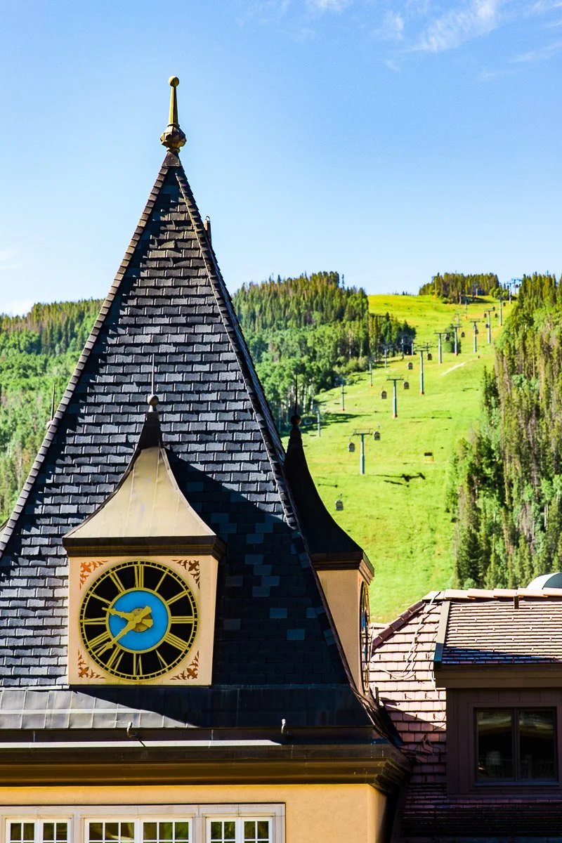 A pointed clock tower with a blue and gold clock face stands in front of a green hillside near ski lift chairs, creating a picturesque scene perfect for a Sonnenalp Hotel wedding under the clear blue sky.