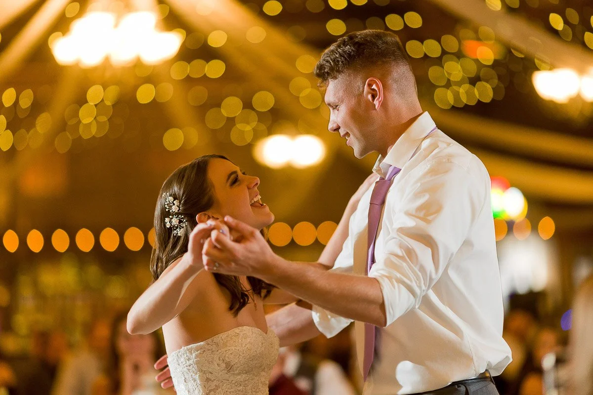 A joyful couple dances at a wedding, the bride in a strapless dress and the groom in a shirt and tie. Warm lights and bokeh create a dreamy atmosphere.