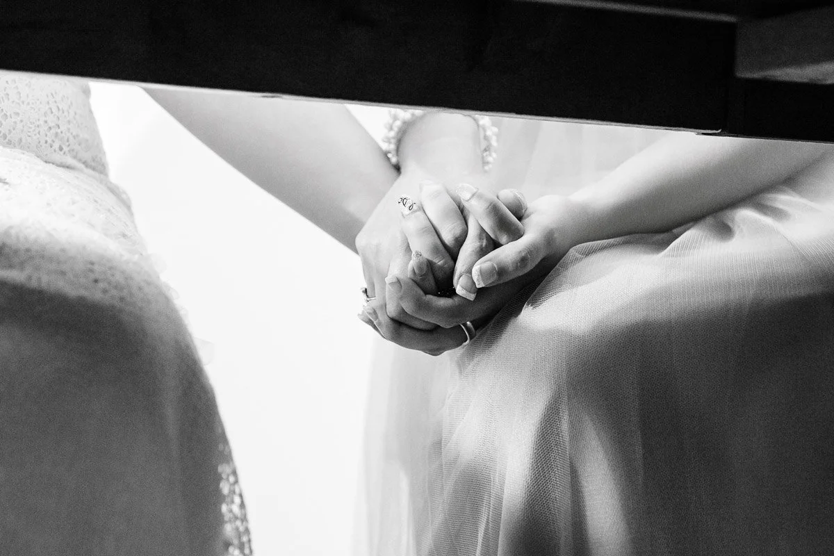 Black and white photo of two people holding hands, both wearing formal dresses. The close-up conveys intimacy and support.