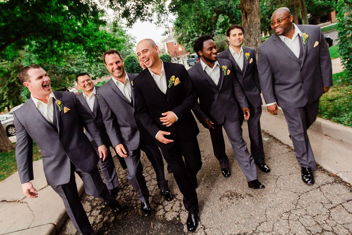Groomsmen in gray suits with yellow boutonnieres walk and laugh down a tree-lined street. Their joyful expressions create a celebratory, cheerful mood.