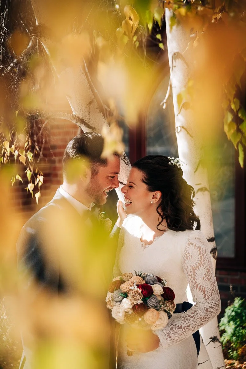 A joyful couple embraces in sunlight under autumn leaves at a Wellshire Event Center wedding, with the bride holding a bouquet of roses. The image conveys warmth and happiness.