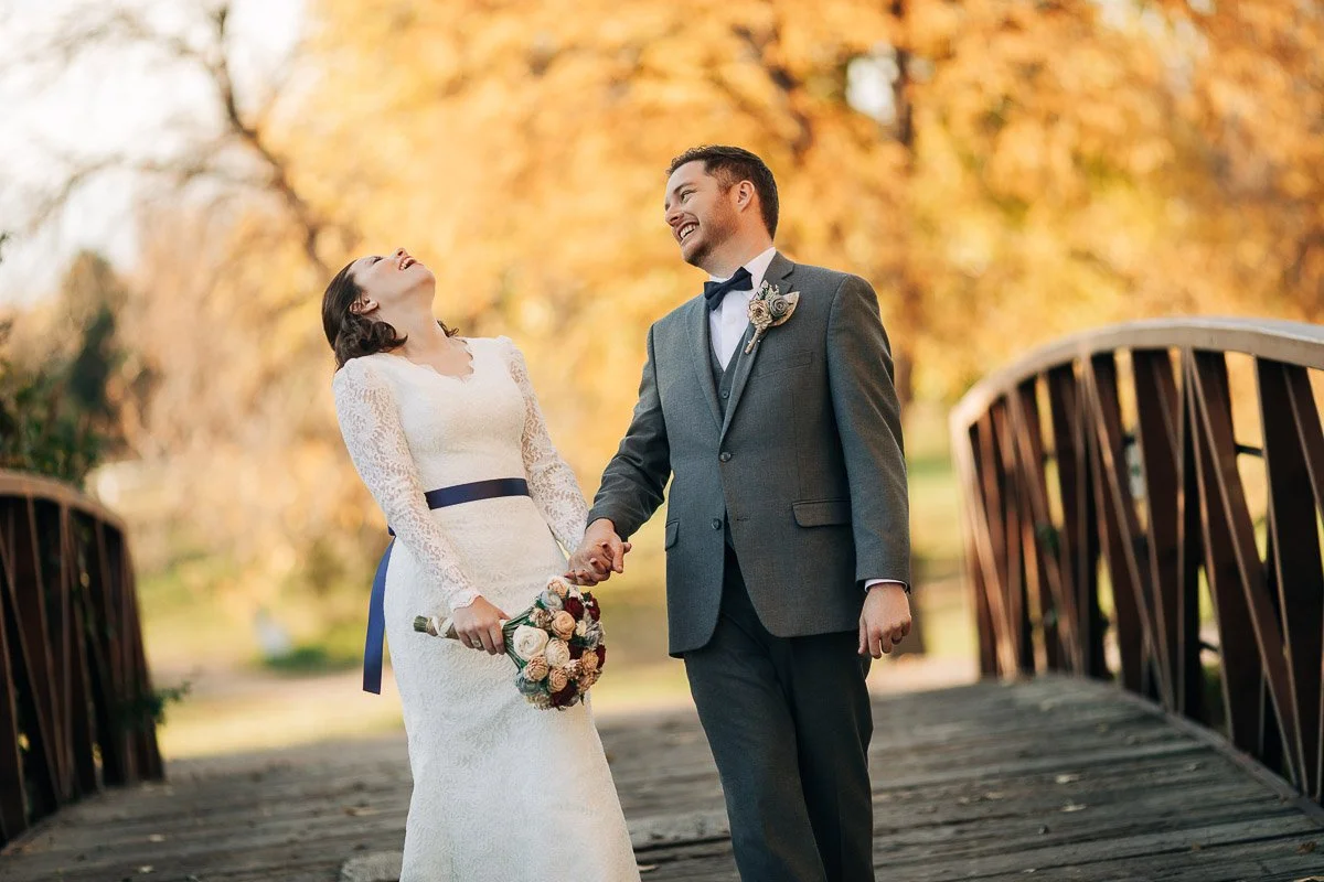 Bride and groom laughing joyfully on a rustic wooden bridge, surrounded by vibrant autumn leaves. She holds a bouquet, wearing a lace gown.