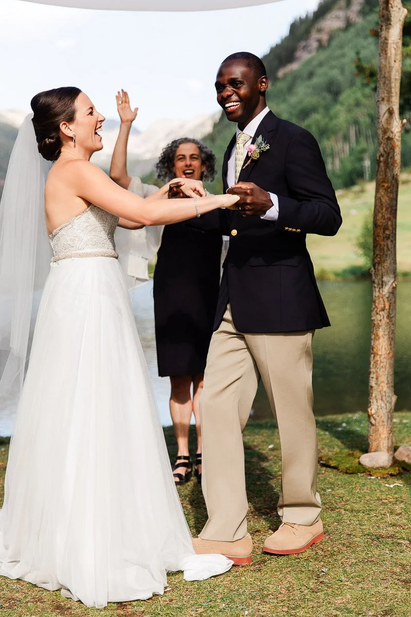 A joyful bride and groom smiling and holding hands during an outdoor wedding ceremony beside a lake with a forested mountain backdrop.