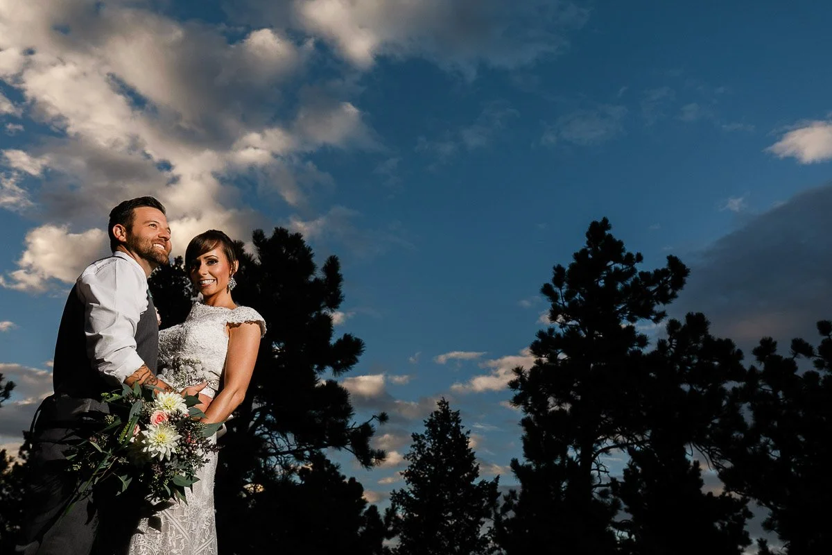 A couple in wedding attire smiles against a vibrant blue sky with soft clouds and silhouetted trees, conveying joy and serenity.