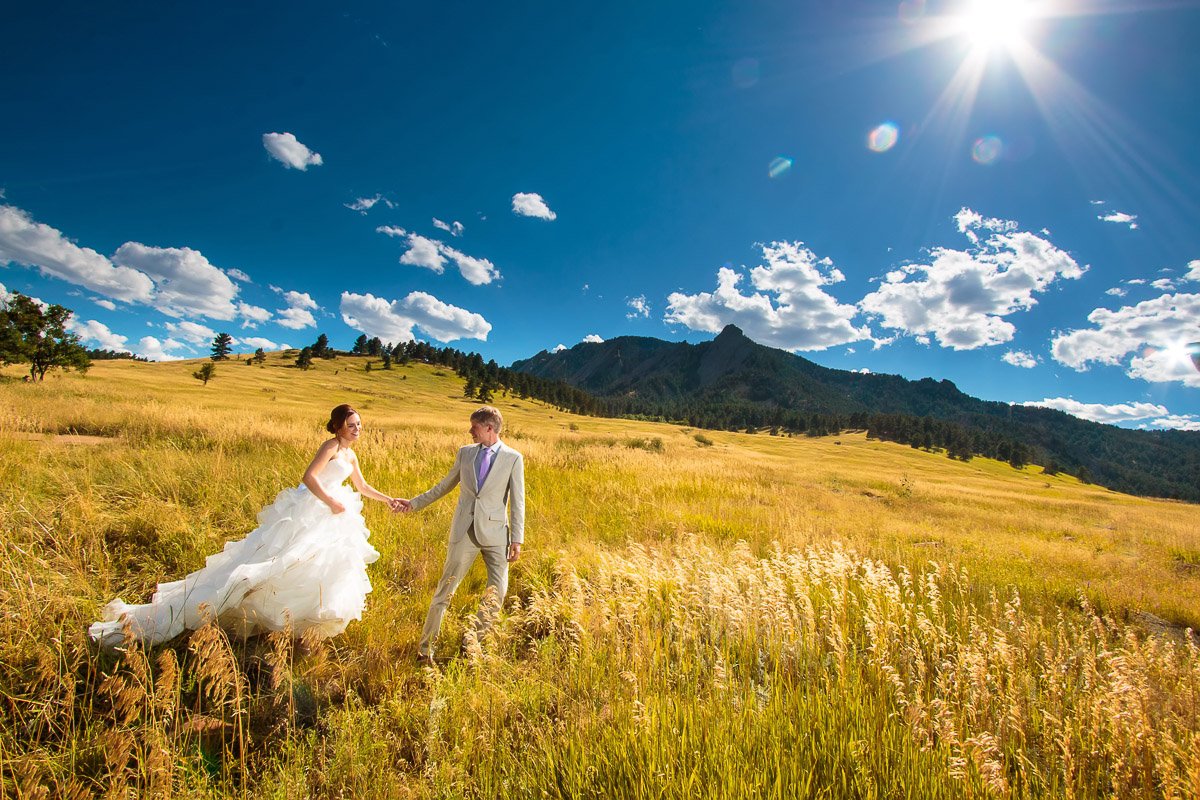 A couple in wedding attire strolls through a sunlit grassy field with mountains and a clear blue sky in the background, evoking a joyful, serene atmosphere.