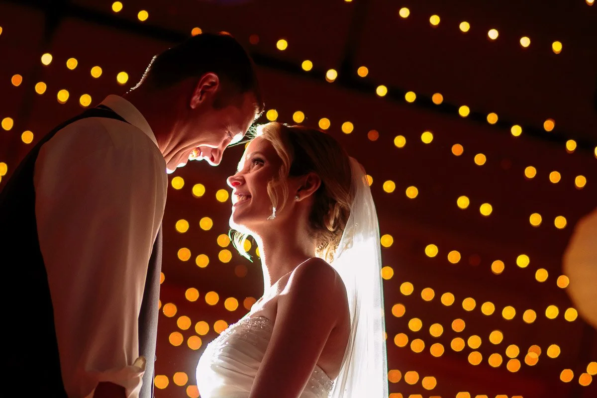 A bride and groom stand close, smiling at each other under warm, glowing string lights during a Silverthorne Pavilion wedding. The setting is intimate and romantic, highlighting their happiness.